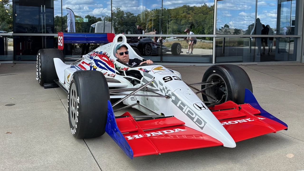 Man sitting in cockpit of IndyCar, pantomiming driving.