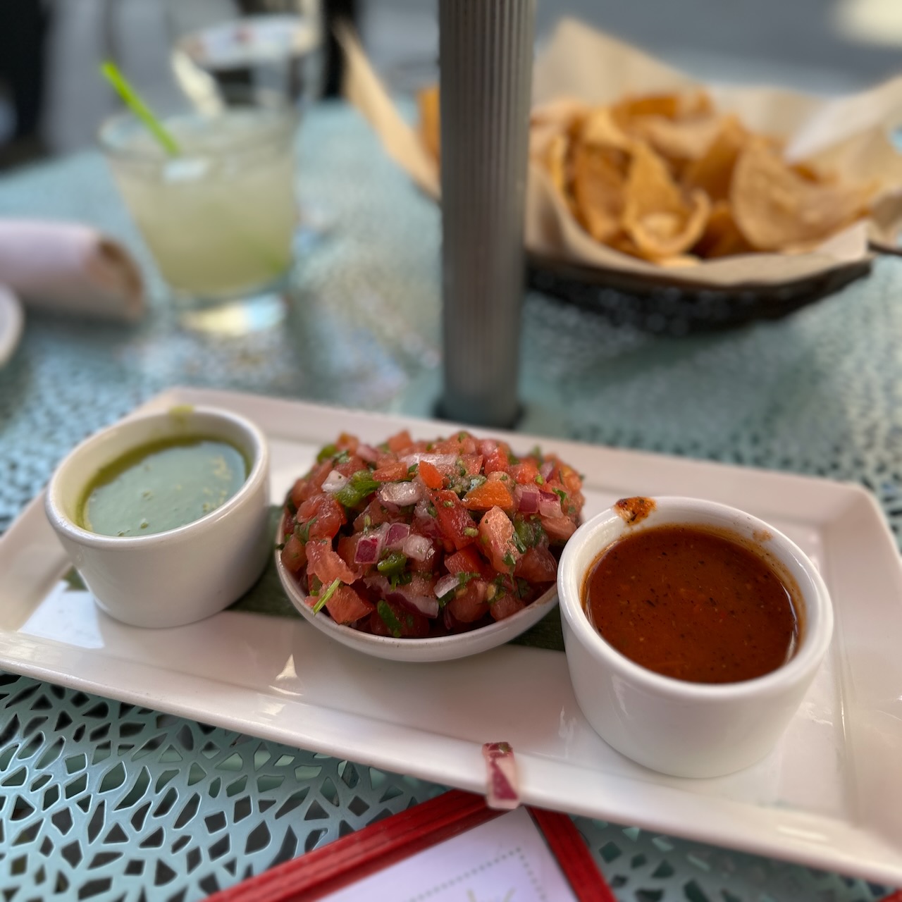 Three different types of salsa, each in a separate cup, on a white plate, with a basket of tortillas chips in the background.