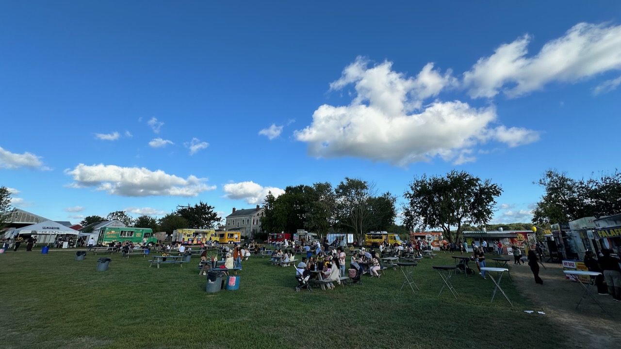 Numerous food trucks in ring around field, with people dining at picnic table in field.