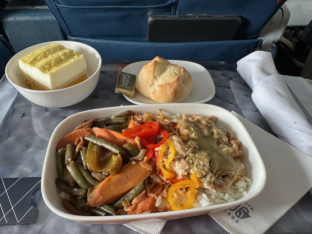 Meal in plastic tray with side dishes of dessert and a roll, all on seat back tray table on airplane.