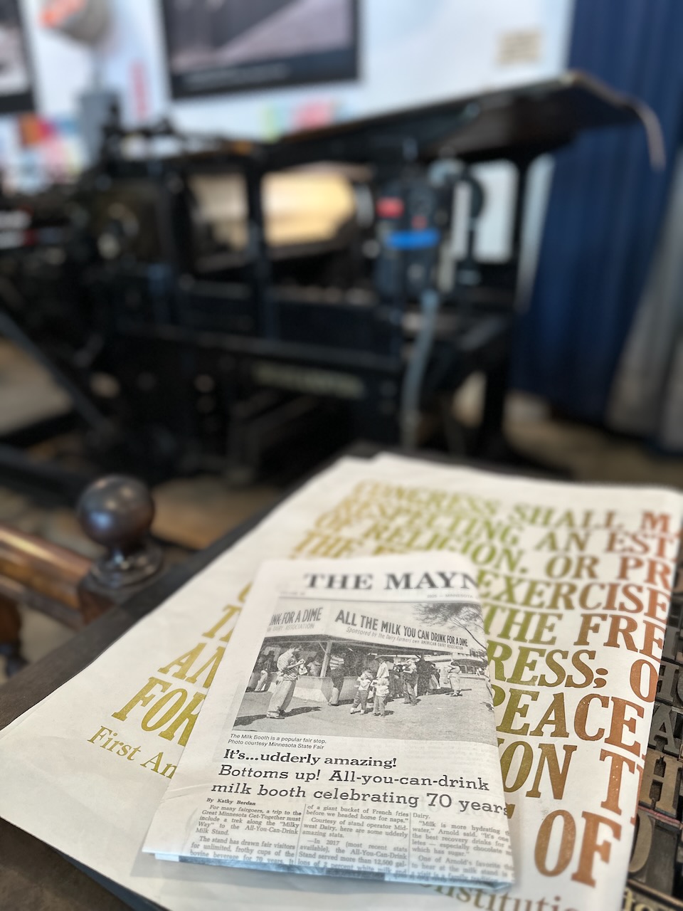 Newspaper on table with old-fashioned printing press in background. 