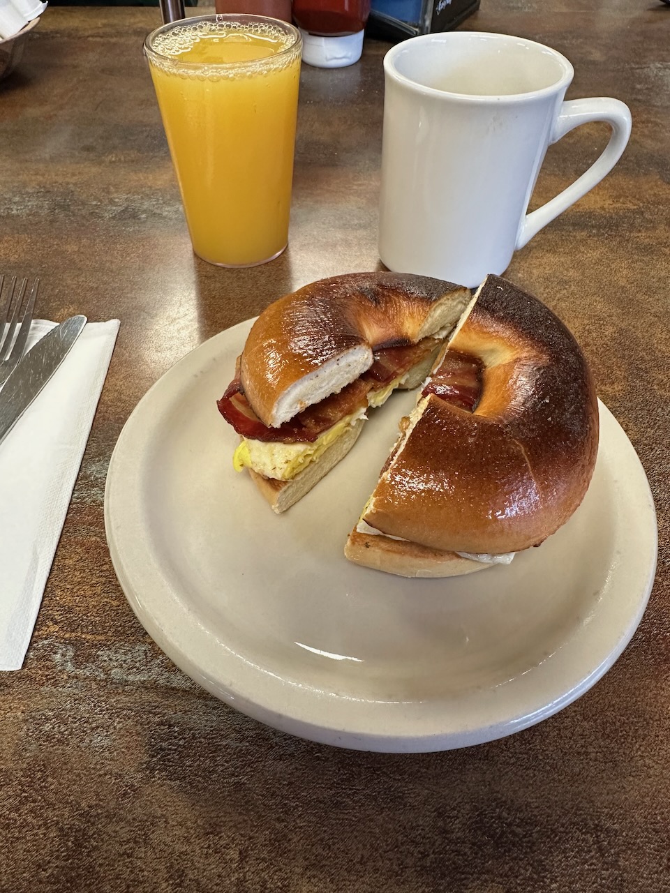 Egg sandwich on bagel on white plate, with orange juice and coffee in cups beside it.