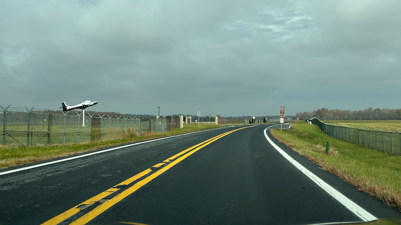 Two-lane road with field on either side of road. A chain link fence is on left side of road with an airplane on pedestal behind the fence. 