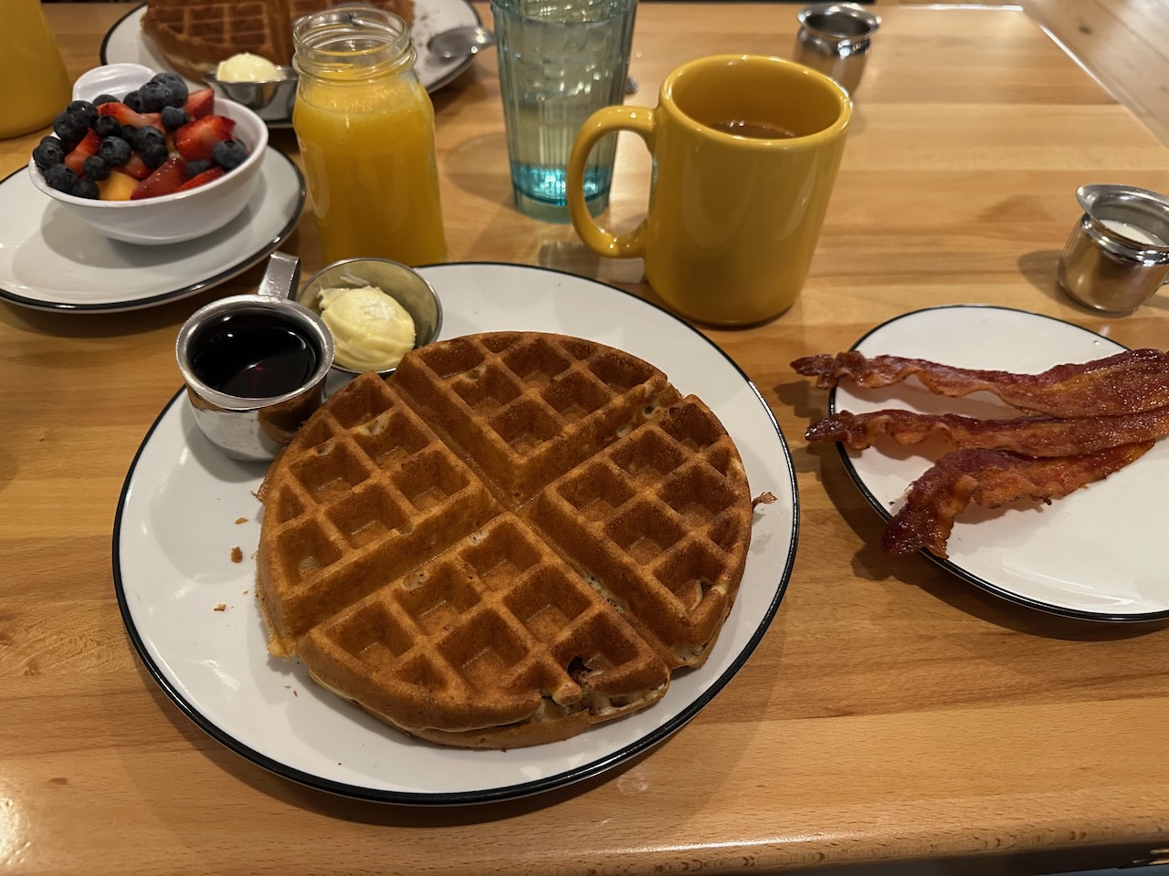 Waffle on plate, with side of bacon, side of fruit, glass of orange juice, and cup of coffee. 