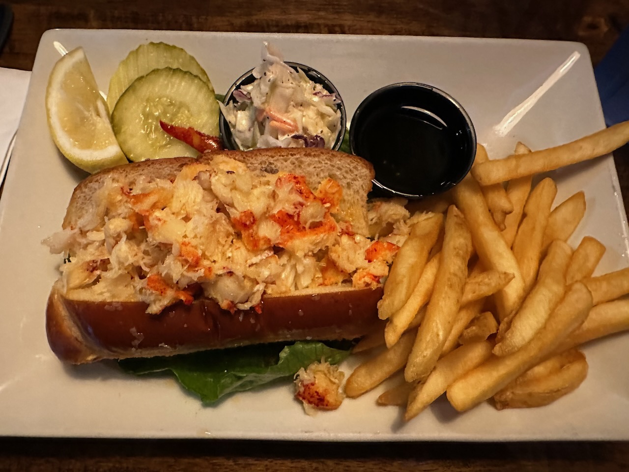 Lobster roll and french fries on white plate, with sides of pickles and coleslaw.