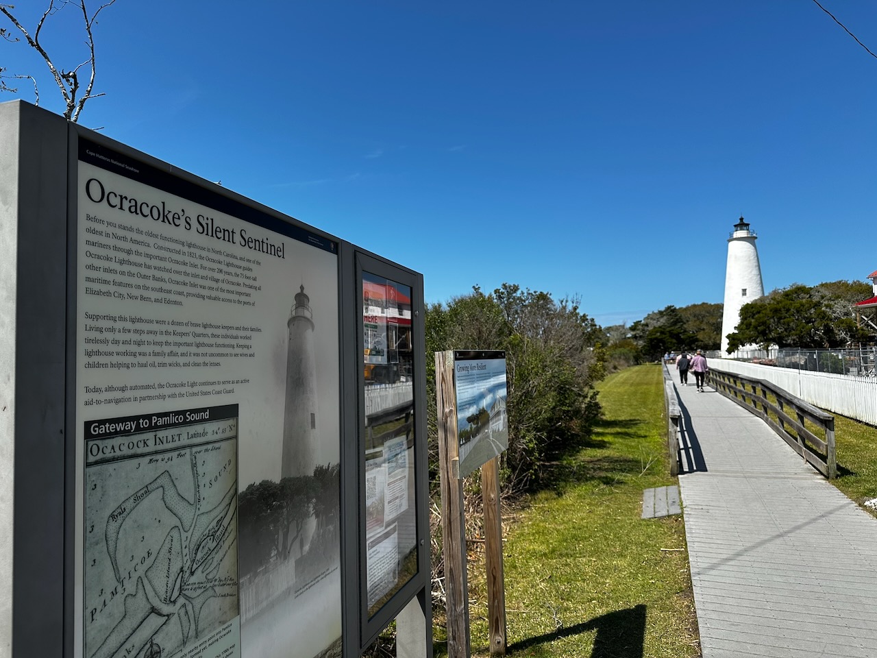 Welcome sign for Ocracoke Ligthouse, with lighthouse in distance at end of walkway.