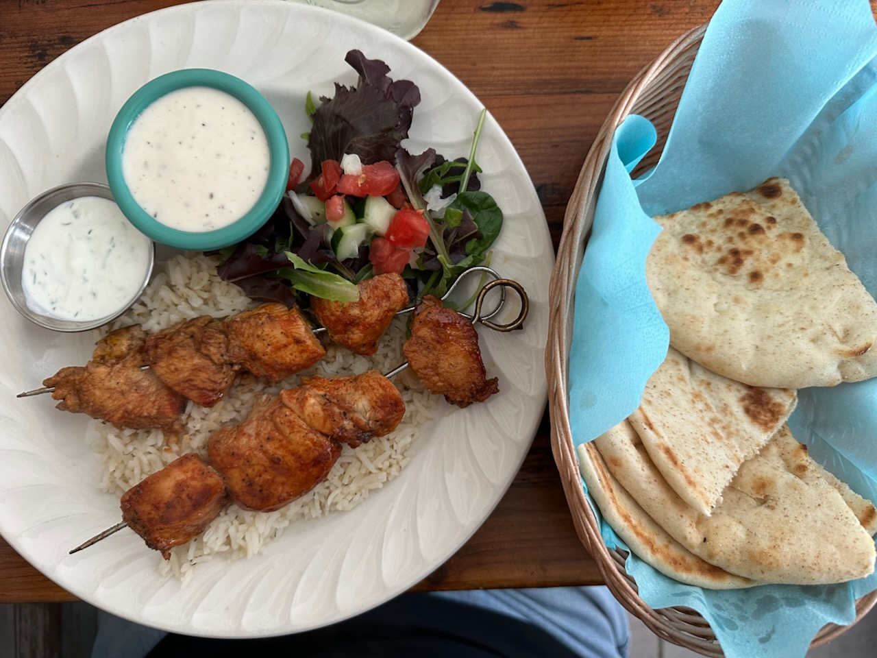 Chicken kabob, rice, salad, and sauces on white plate, with basket with pita bread on side.