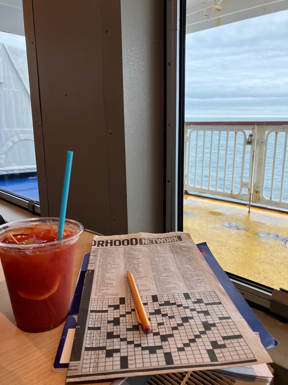 Crossword puzzle and Bloody Mary drink in cup, both on wooden table with view out window of ferry.
